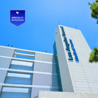 Toma desde un ángulo bajo del edificio del Centro Médico General de Los Ángeles contra un cielo azul despejado, con una insignia azul de Excelencia en Especialidades en la esquina superior izquierda.
