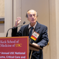 Close-up of Dr. Demetrios Demetriades speaking at a podium during the symposium, gesturing with his hand.
