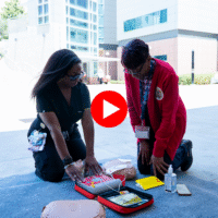 Two women kneeling outdoors during a community Sidewalk CPR training session, practicing with an AED training device and mannequin parts.