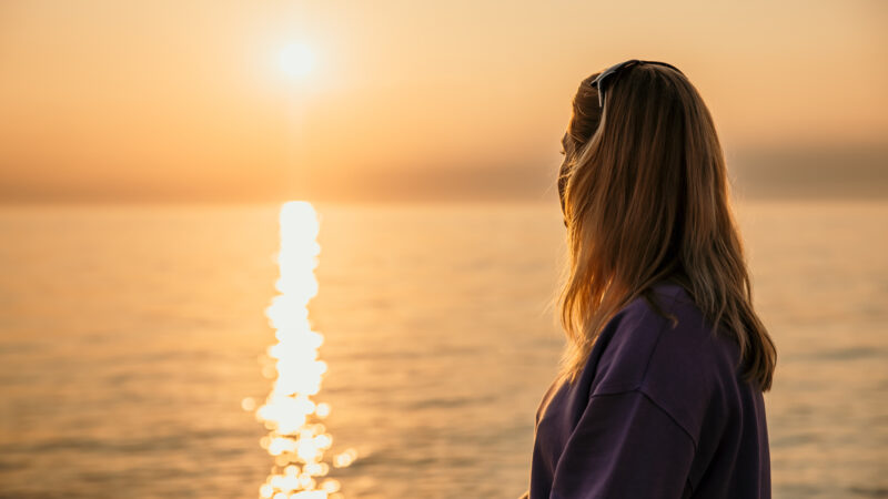 Mujer adulta con cabello rubio disfrutando de la puesta de sol en el mar con reflejo en la superficie del agua